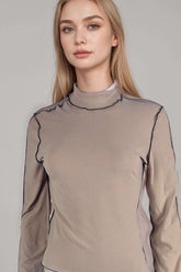 Woman wearing sheer beige mock neck long sleeve top with contrast black stitching
